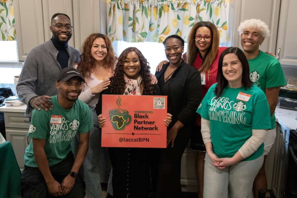 A multiracial group of people smile, including Starbucks employees. The woman in the center holds a sign that reads "Starbucks Black Partner Network"