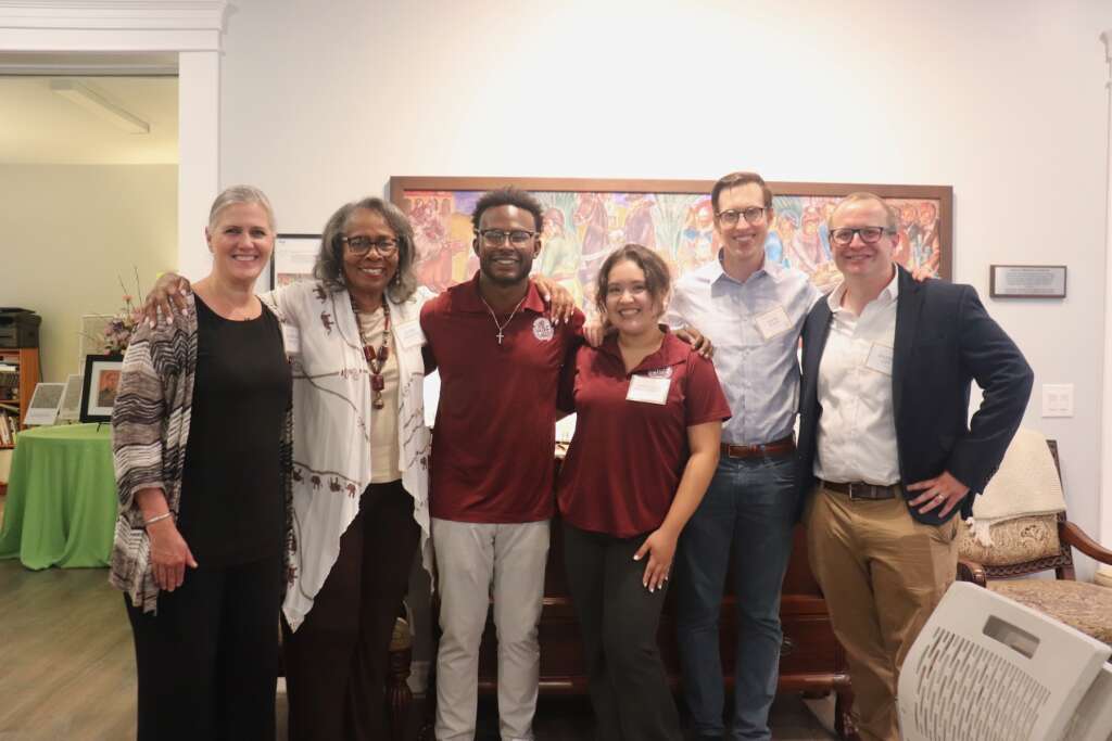 A multiracial group of people stand with their arms around each other